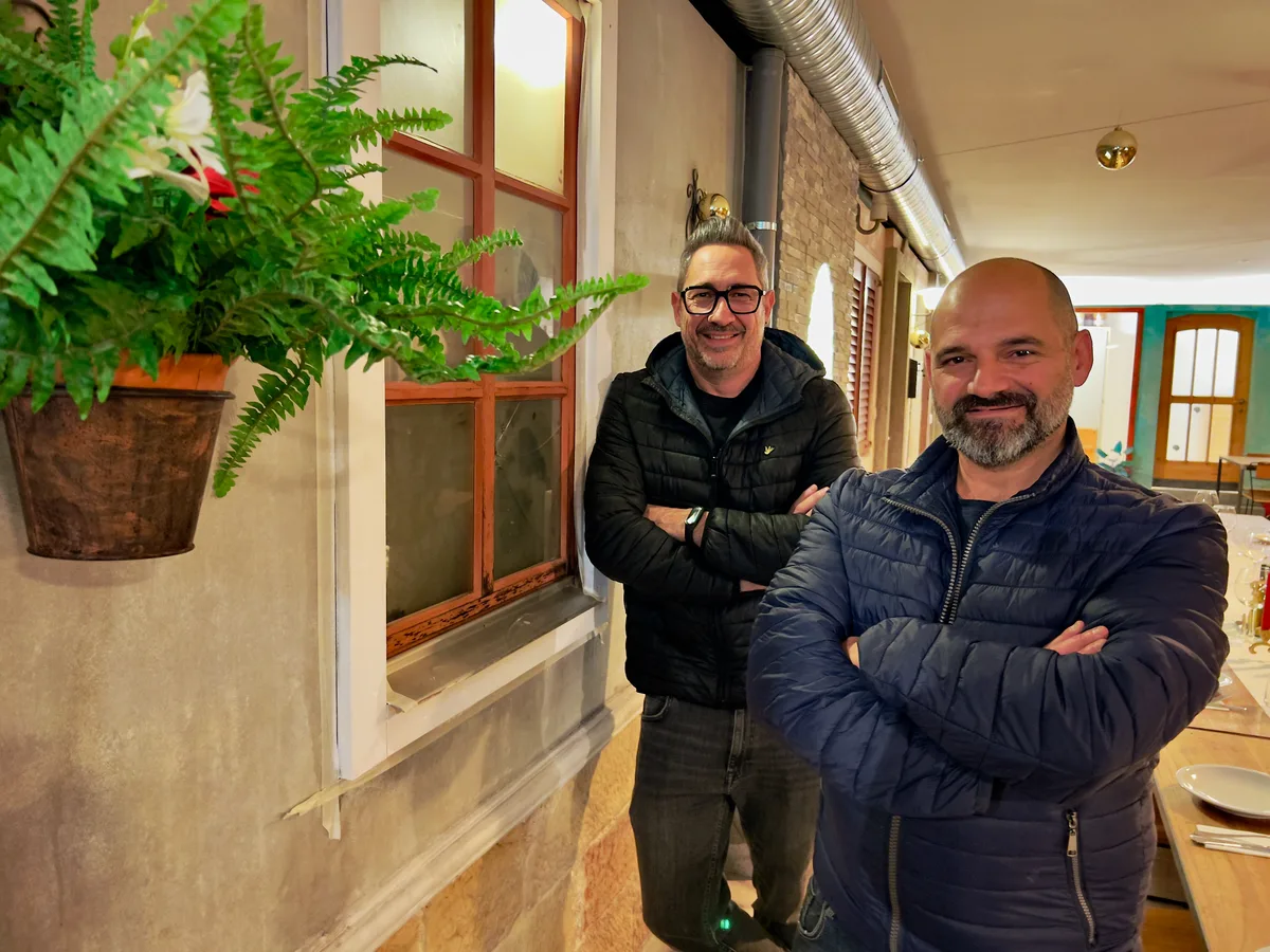 Zwei Männer stehen mit verschränkten Armen neben einer Wand, die an eine Häuserfassade mit Fenstern und Türen erinnert. Beide lächeln in die Kamera.